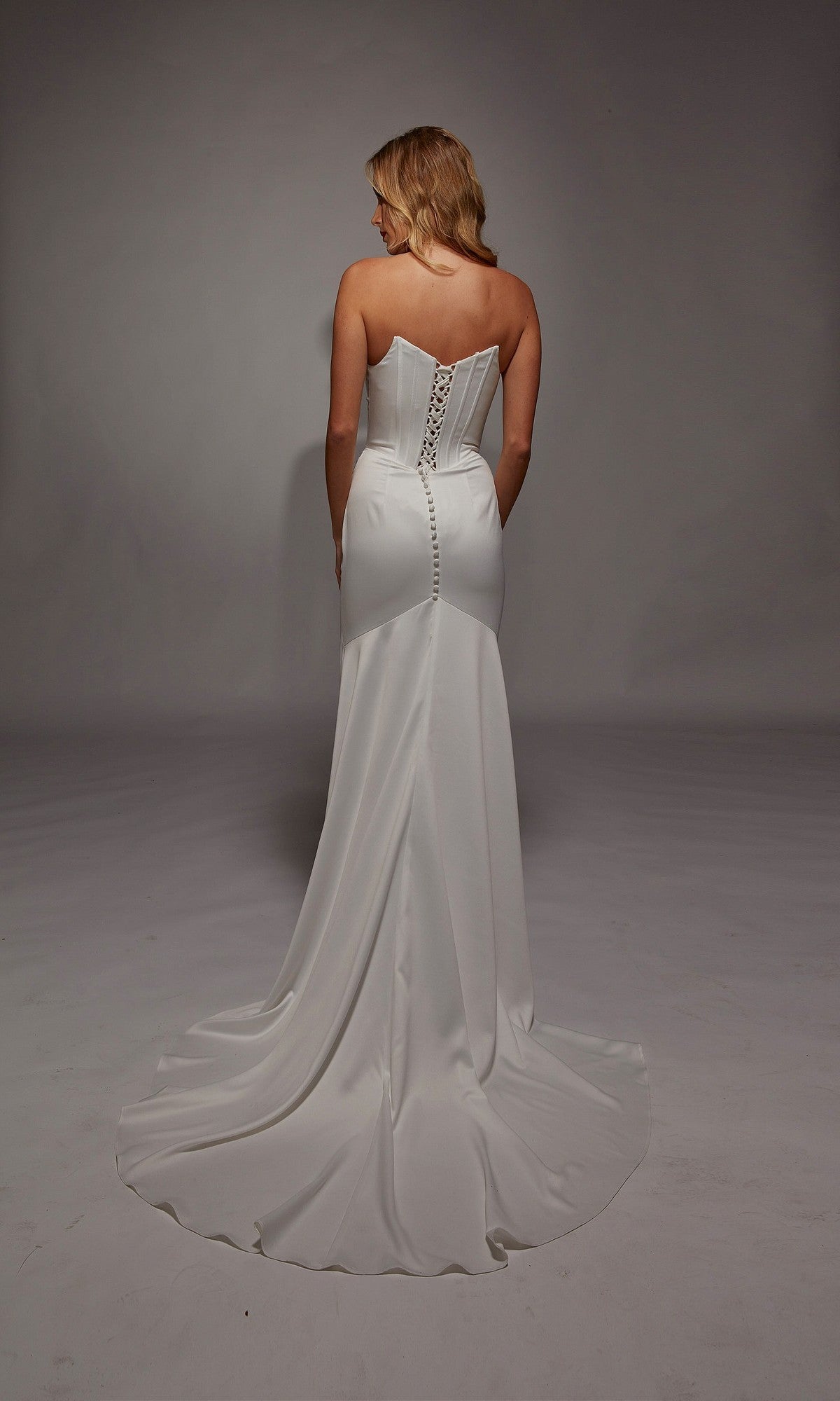 A woman stands facing away against a plain background, wearing the Long White Dress: Alyce 7139—a strapless satin gown with a lace-up back and elegant button details.